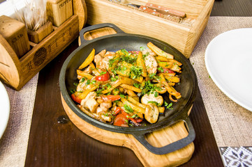 fried potatoes with mushrooms, meat and greens on a black frying pan on a wooden table in a restaurant, next to 2 white plates, appliances and spices