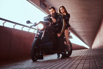 Fototapeta na wymiar Brutal bearded biker in black leather jacket and sensual brunette girl sitting together on a custom-made retro motorcycle on footway under a bridge.