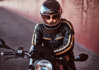 Close-up portrait of a brutal bearded biker in helmet and sunglasses dressed in a black leather jacket sitting on a retro motorcycle with an included headlight.
