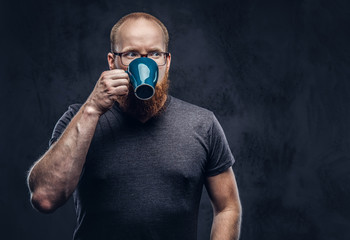 Close up portrait of a redhead bearded male drinks coffee wearing glasses dressed in a gray t-shirt, isolated over a dark textured background.