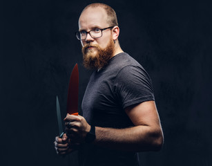 Portrait of a redhead bearded male wearing glasses dressed in a gray t-shirt, standing with colored steel knives in his hands. Isolated on a dark textured background.