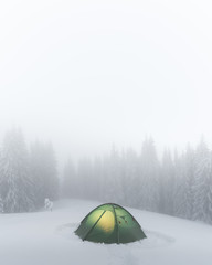 Green tent lighted from the inside by a flashlight against the backdrop of foggy pine tree forest. Amazing snowy landscape. Tourists camp in winter mountains. Travel concept