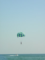 The man was attached to a long line to a moving boat. On a blue-green parachute, a man soars in the air. Theme active recreation in the resort, parasailing, having fun in summer