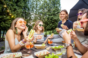 Young friends having fun sitting together with tasty snacks and drinks during the evening lights at...