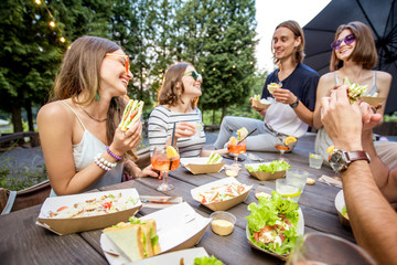 Young friends having fun sitting together with tasty snacks and drinks during the evening lights at the park cafe outdoors