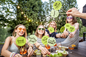 Young friends holding green plates with healthy food slogans sitting with tasty food at the outdoor...