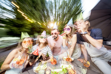 Happy friends having fun together celebrating birthday at the outdoor park cafe during the evening light. Motion blur image technic