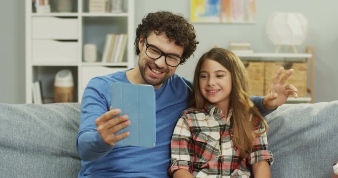 Portrait of the cheerful dad and small daughter sitting on the sofa and having a nice videochat on the tablet device. Indoors.