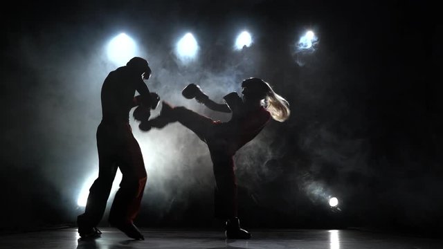 Girl is kicking a guy with their sparring kickboxing. Light from behind. Smoke background. Silhouette