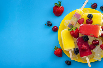 Summer treats and sweet frozen desserts concept with assorted popsicles and berry fruits like strawberry, cherry, blackberry and raspberry isolated on minimalist blue background with copy space