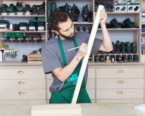 The carpenter bearded hipper in working clothes works in the workshop with wooden details.