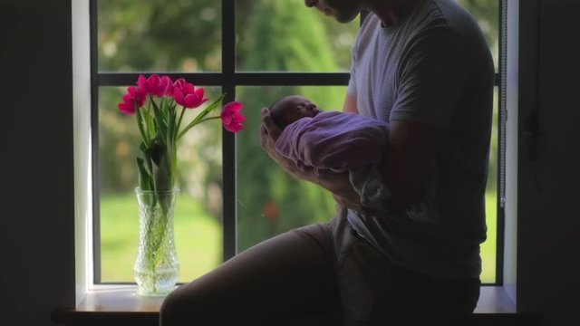 Father cradling swaddled newborn baby in arms. Man rocking newborn baby by window.