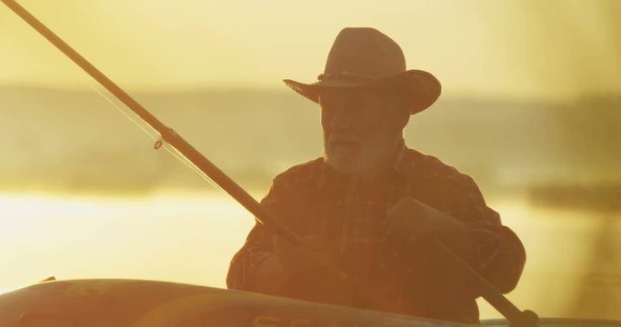 Close up of the senior fisherman in the hat floating in the boat and fishing with a rod in hands in the dawn. Outside.