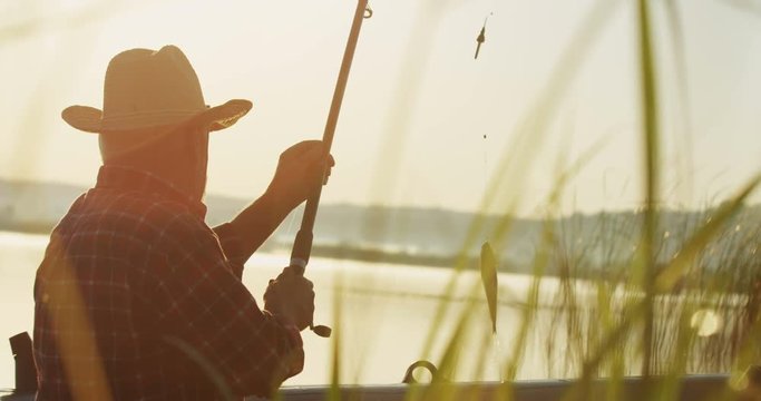 Back view on the old fisherman in a hat catching a fish on a rod at the dawn on a lake. Outdoor.