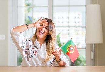 Young woman at home holding flag of Portugal with happy face smiling doing ok sign with hand on eye looking through fingers