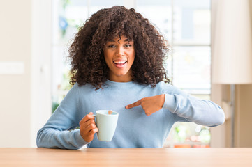 African american woman holding a cup of coffee at home with surprise face pointing finger to himself