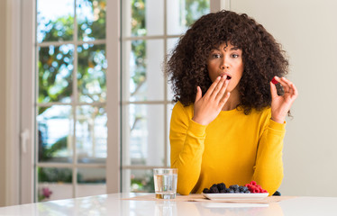 African american woman eating raspberries and blueberries at home cover mouth with hand shocked...