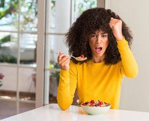 Fototapeta na wymiar African american woman eating cereals, raspberries and blueberries annoyed and frustrated shouting with anger, crazy and yelling with raised hand, anger concept