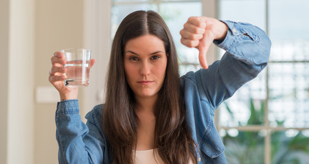 Young woman drinking glass of water at home with angry face, negative sign showing dislike with...