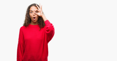 Obraz na płótnie Canvas Young hispanic woman wearing red sweater doing ok gesture shocked with surprised face, eye looking through fingers. Unbelieving expression.