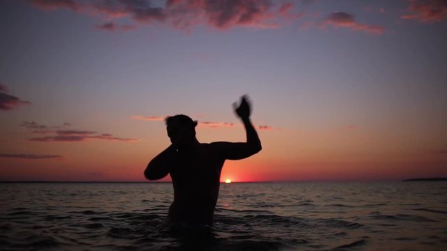 Silhouette of happy joyful man playing around and dancing in water, after swimming in sea during beautiful sunset in slow motion