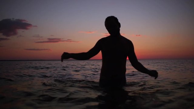 Silhouette of cheerful strong man taking pleasure and dancing in water, after swimming in sea during beautiful sunset in slow motion