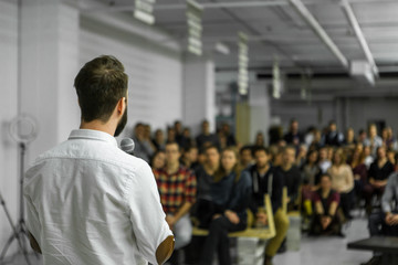 Man with beard gives a public speech in front of 200 people, in an industrial environment