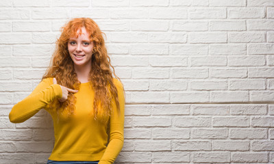 Young redhead woman standing over brick wall with surprise face pointing finger to himself