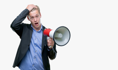 Young handsome blond man holding a megaphone stressed with hand on head, shocked with shame and surprise face, angry and frustrated. Fear and upset for mistake.