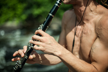Topless muscular man plays clarinet with a blurry nature view in the background - obrazy, fototapety, plakaty