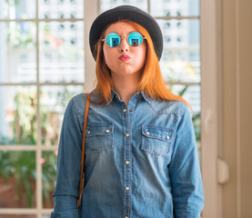 Stylish redhead woman wearing bowler hat and sunglasses puffing cheeks with funny face. Mouth...