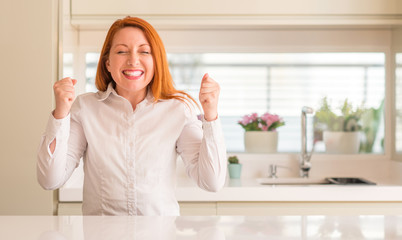 Redhead woman at kitchen excited for success with arms raised celebrating victory smiling. Winner concept.