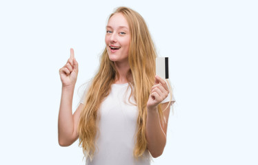 Blonde teenager woman holding credit card surprised with an idea or question pointing finger with...