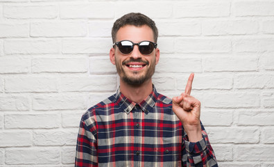 Young adult man wearing sunglasses standing over white brick wall surprised with an idea or...