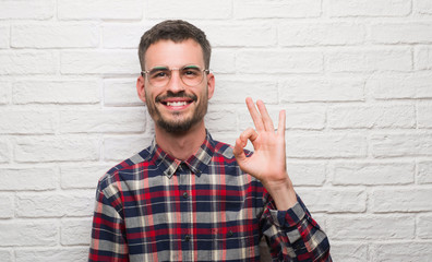 Young adult man standing over white brick wall doing ok sign with fingers, excellent symbol
