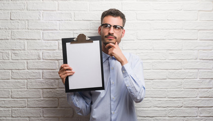 Young adult business man over brick wall holding clipboard serious face thinking about question,...