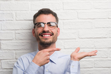 Young adult business man standing over white brick wall very happy pointing with hand and finger