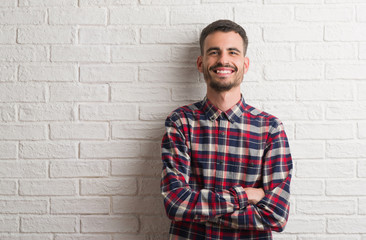 Young adult man standing over white brick wall happy face smiling with crossed arms looking at the...