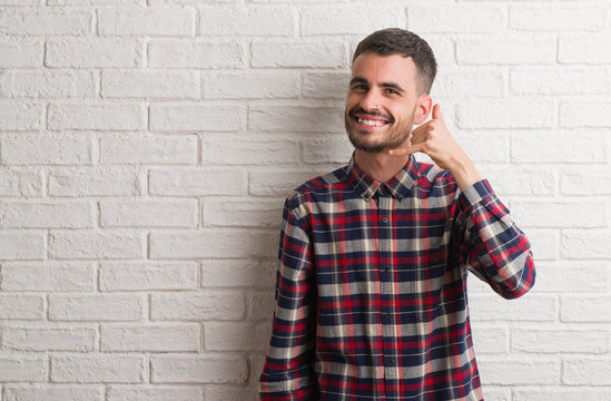 Young adult man standing over white brick wall smiling doing phone gesture with hand and fingers like talking on the telephone. Communicating concepts.