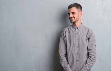 Young adult man standing over grey grunge wall looking away to side with smile on face, natural expression. Laughing confident.