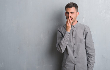 Young adult man standing over grey grunge wall bored yawning tired covering mouth with hand. Restless and sleepiness.