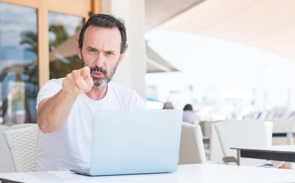 Handsome senior man using laptop at restaurant pointing with finger to the camera and to you, hand sign, positive and confident gesture from the front