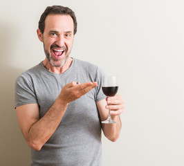 Senior man drinking red wine in a glass very happy pointing with hand and finger