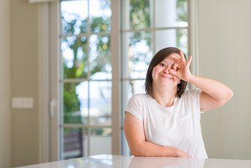 Down syndrome woman at home doing ok gesture with hand smiling, eye looking through fingers with happy face.