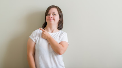 Fototapeta premium Down syndrome woman standing over wall cheerful with a smile of face pointing with hand and finger up to the side with happy and natural expression on face looking at the camera.