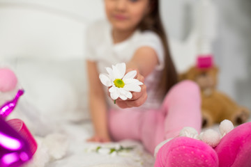Obraz na płótnie Canvas Cute little flower in hands of preschooler girl. Close up picture of pretty white daisy flower being held by little girl that is sitting on white blanket.