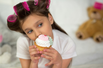 Adorable little girl eating fresh sweet cupcake indoors. Sweet little preschooler tilting her head to take bite of white iced cupcake.