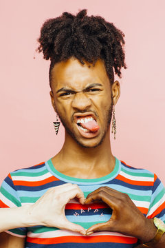 Portrait of young man with earrings sticking tongue out, with happy heart shaped with hands on his shirt
