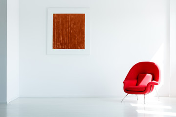Red painting on white wall in spacious empty loft interior with modern armchair. Real photo