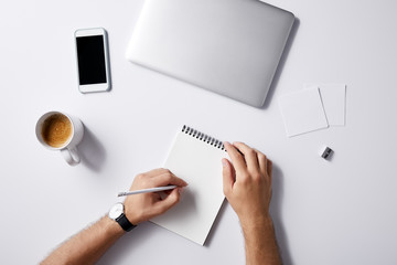 cropped shot of man writing in notebook at workplace with gadgets and coffee cup on white surface for mockup